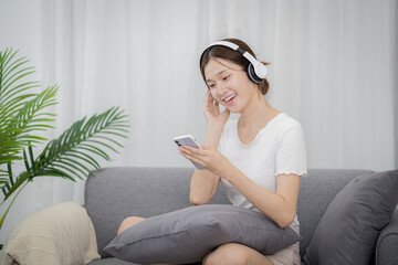Obraz premium Relaxed young Beautiful Asian woman music lover listening music on earphones holding mobile phone in living room listening music with headphones and mobile phone