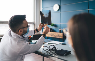 Male doctor in mask and glasses examining x ray image with vertebra model
