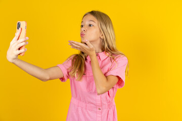 Beautiful caucasian kid girl wearing pink shirt blows air kiss at camera of smartphone and takes selfie, sends mwah via online call.