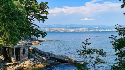 Fototapeta na wymiar A panoramic view of the shore along Lovran, Croatia. There is a small town located between the lush trees. The Mediterranean Sea has turquoise color. Big boulders in the water. Sunny day. Calmness