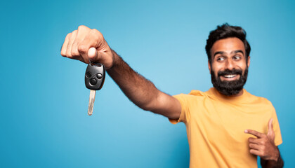 New car. Happy indian man showing auto key and pointing at it in excitement, posing on blue background, selective focus