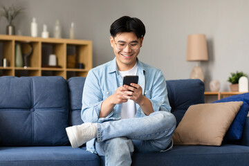 Obraz premium Happy Korean Young Man Using Smartphone Texting Sitting At Home