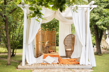 Rattan peacock armchair and decorative folding screen divider. Summer gazebo with flowing white curtains. Wedding boho decoration. Decor outdoor terrace with wicker furniture. Outdoor design of arbour © stock_studio