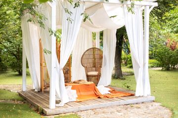 Outdoor design of arbour. Rattan peacock armchair and decorative folding screen divider. Summer gazebo with flowing white curtains. Wedding boho decoration. Decor outdoor terrace with wicker furniture