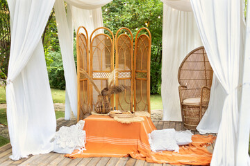 Summer gazebo with flowing white curtains. Wedding boho decoration. Decor outdoor terrace with wicker furniture. Outdoor design of arbour. Rattan peacock armchair and decorative folding screen divider © stock_studio