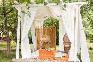 Summer gazebo with flowing white curtains. Wedding boho decoration. Decor outdoor terrace with wicker furniture. Outdoor design of arbour. Rattan peacock armchair and decorative folding screen divider © stock_studio
