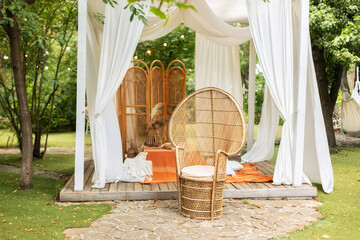 Rattan peacock armchair and decorative folding screen divider. Summer gazebo with flowing white curtains. Wedding boho decoration. Decor outdoor terrace with wicker furniture. Outdoor design of arbour © stock_studio
