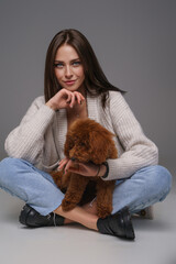 Chic brunette in a white top and denim shorts sits cross-legged, holding her brown toy poodle against a gray studio backdrop