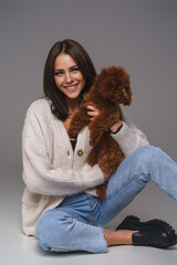 Stunning brunette in casual, sitting on the floor, intimately holding her toy poodle, framed by a serene gray studio setting