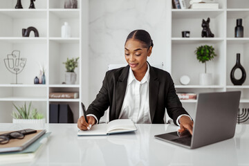 Cheerful black businesswoman with laptop taking notes, writing business ideas or daily schedule,...