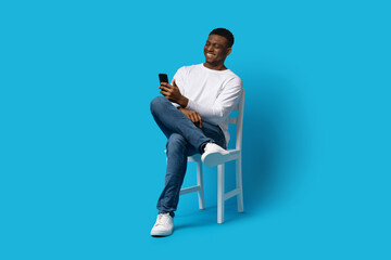 Happy young black man sitting on chair, using cell phone