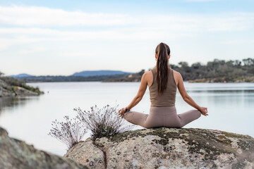 Fototapeta na wymiar Beautiful woman doing relaxation and meditation exercises in nature. Enjoying and appreciating the beauty of nature that surrounds you on top of a rock on the shores of a lake