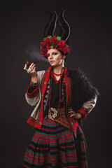 beautiful brunette woman in a traditional Ukrainian costume, with a floral wreath and horns, is standing on the black studio background and smoking a pipe