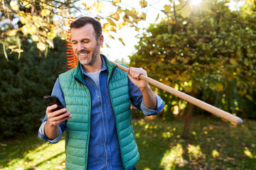 Man using his mobile phone in garden standing with leaf rakes during sunny autumn afternoon
