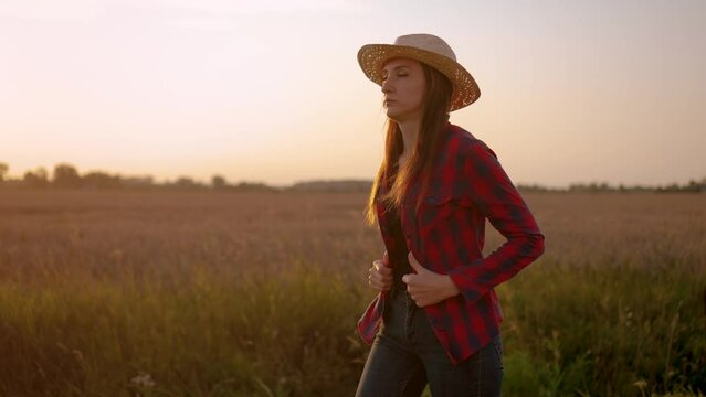 Farmer woman in red shirt and straw hat walking through ripe wheat field at sunset. Work in agronomic farm business and production organic eco bio food