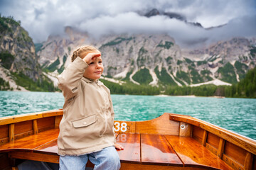 Little girl sitting in big brown boat at Lago di Braies lake in cloudy day, Italy. Summer vacation in Europe