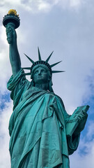 A close up on an iconic representation of freedom and independence, the Statue of Liberty with flaming torch on Liberty Island. The Lady on a Pedestal is surrounded by clouds. American history.