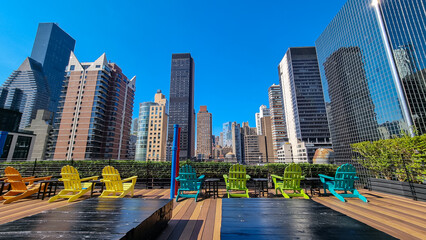 A rooftop with the city skyline view and modern skyscrapers against a sunny blue sky in New York. Colorful chairs overlooking the city. Rows of skyscrapers in the back. Sunny and warm day.