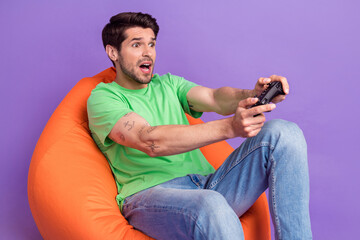 Photo of young funny staring man hold playstation gamepad console sitting pouf addicted losing isolated on violet color background