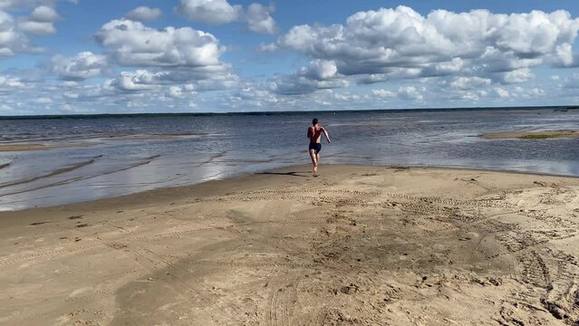 sportsman on the beach makes jumps to warm up and runs into the water. a teenager on the seashore performs physical exercise.
