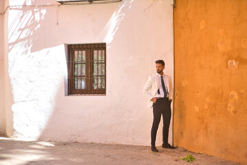 Attractive young businessman with beard, suit and tie, posing with his hands in his pockets next to two white and orange walls. Concept beauty, fashion, success, achiever, trend.