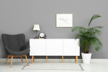 Interior of stylish living room with drawers, armchair, houseplant and blank frame hanging on grey wall