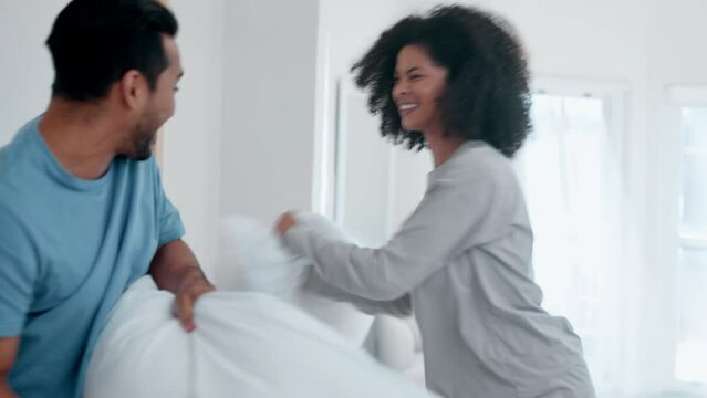 Happy couple, bed and playing pillow fight in fun morning, wake up or bonding together at home. Interracial man and woman smile in happiness for weekend, holiday or break with cushion in bedroom