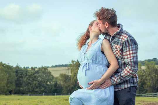 inniger Kuss einer hochschwangeren jungen blonden Frau mit dickem Babybauch und ihres blond gelockten Mannes vor hügeliger Landschaft im Sommer