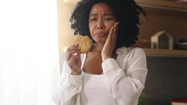 Portrait of young curly woman eating cookie and having toothache standing at kitchen at home Sad female feeling pain in tooth because caries gum disease or sensitive teeth indoors