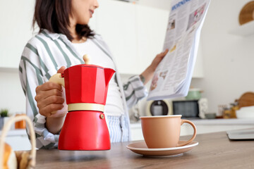 Beautiful young Asian woman with geyser coffee maker and cup of espresso reading newspaper in kitchen