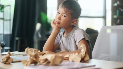 Portrait of exhausted school boy with failed crumped papers while sitting with head on hand at home Tired little kid child with creative block or lack of inspiration indoors Stress education concept