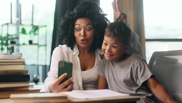Cute funny young curly woman mother with little son kid and grimacing taking selfie photograph saving great memories on smartphone mobile phone while take break of studying at home Happy motherhood