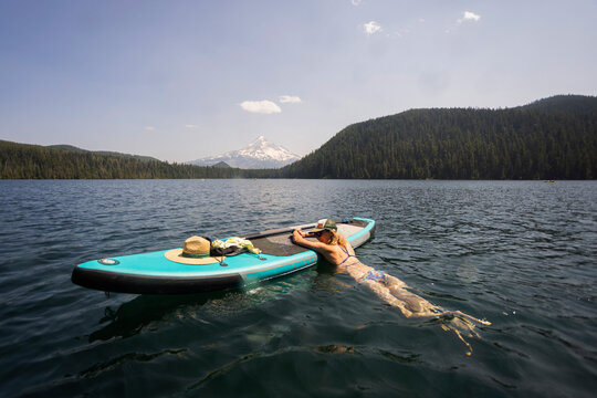 A young woman cools off in an alpine lake at the base of Mt. Hood with her standup paddle board on a summer day. 