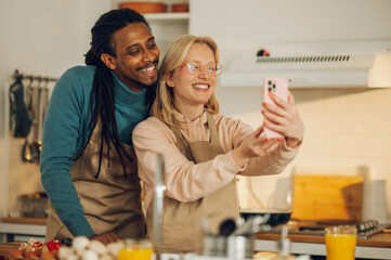 A happy multicultural couple is taking self-portraits in the kitchen while cooking dinner at home together.