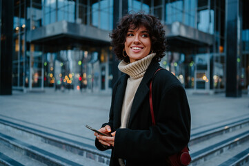 Young woman holding her smart phone in her hands and looking away with a smile.