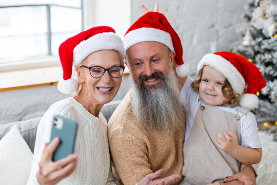 Senior grandparents, attractive grandmother and handsome grandfather taking photo selfie with cute little granddaughter near decorated Christmas tree. Two generations, family values, happy people
