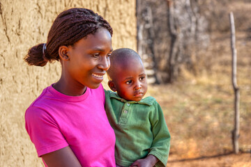 African mother holding her son, sited in front of her home, in the sandy yard, in a late afternoon...