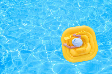 Baby swim in a yellow inflatable raft in a pool of blue water. Small child swims alone with a shovel in yellow rubber ring. View from the top.