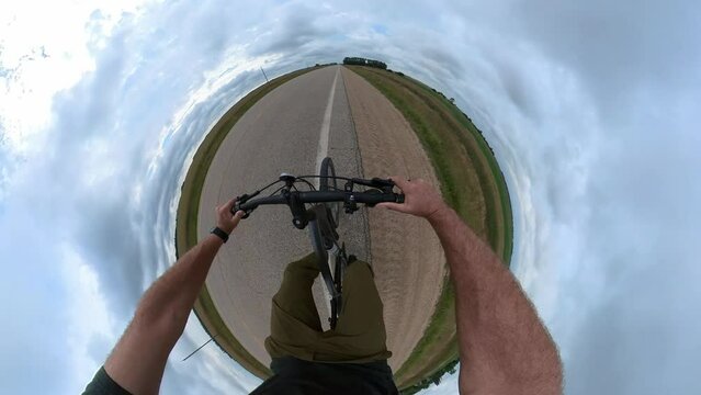 Action camera POV as a 360 degree tiny planet style video of a person riding a bicycle along a paved highway. The view includes the cyclist&rsquo;s feet which are pedaling quickly.