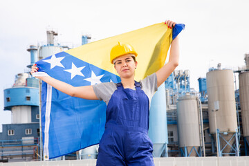 Sad female worker in hardhat with bosnian flag standing in front of factory