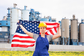 Sad unhappy young female worker in helmet waving national flag of USA during strike in front of big...