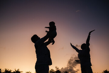 
Silhouettes of the family against the backdrop of the sunset sky, dad holds the baby high above his head, mom joyfully threw her hands up.