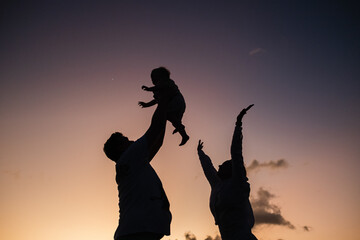 
Silhouettes of the family against the backdrop of the sunset sky, dad holds the baby high above his head, mom joyfully threw her hands up.