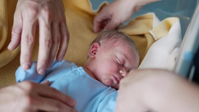 Loving parents stand near their sleeping newborn. Tiny baby boy in blue romper resting caressed by dad's hand. Close up.