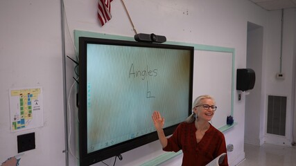 Happy female teacher writing on an interactive whiteboard teaching geometry math in a school classroom with US American flag in background.