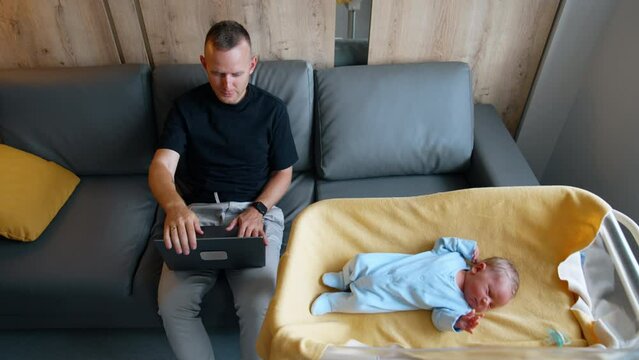 Mid-aged man works on his laptop sitting on the sofa. Father distracts on his newborn baby lying in the crib nearby. Top view.