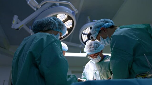 Group of surgeons and nurse stand around the patient on the operational table. Male chief surgeon uses a mallet. Low angle view.