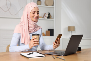 Muslim woman with cup of coffee using smartphone near laptop at wooden table in room