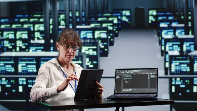 Senior computer scientist in server farm completing tasks on tablet while running scripts on laptop terminal. Expert amidst supercomputers manipulating code and improving cybersecurity
