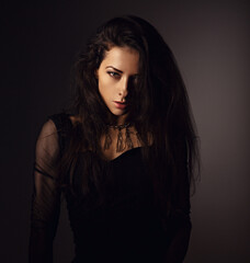 Naklejka premium Beautiful woman in black dress posing in fashion necklace on the neck. Long hairstyle on dark shadow background. Toned portrait. Closeup.